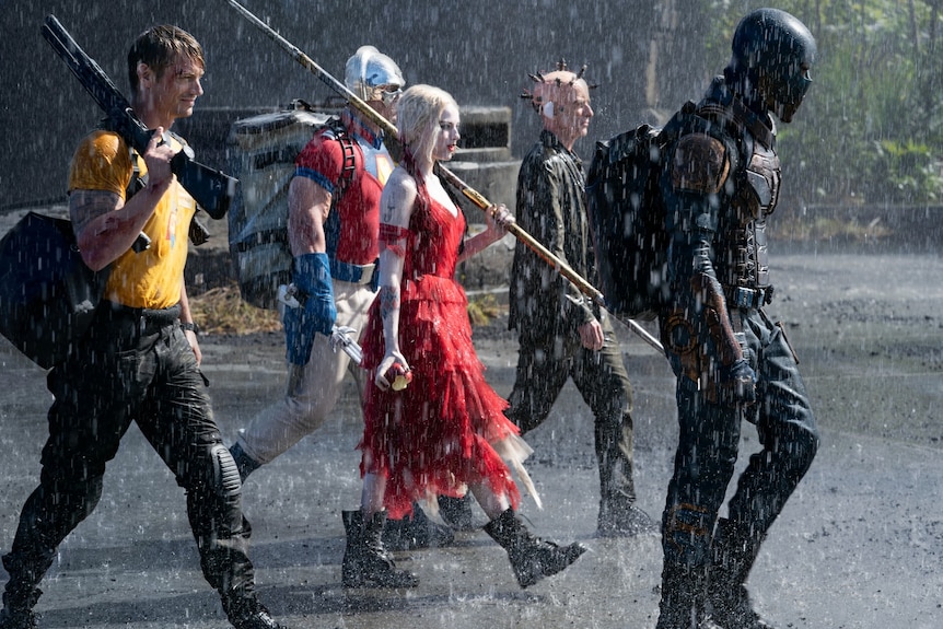 Five people walk in the rain, holding weapons, including superheroes/villains Harley Quinn, The Thinker and Peacemaker