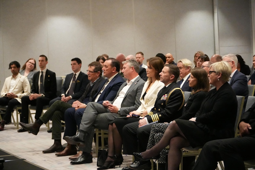 A crowd of well dressed people listen to someone speaking in a room.