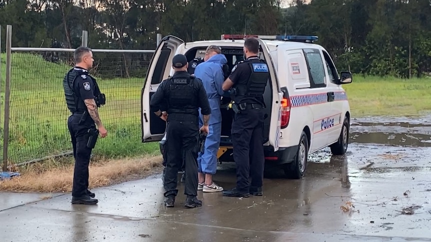 Officers arrest a man dressed in a blue forensic suit at the back of a marked police car.