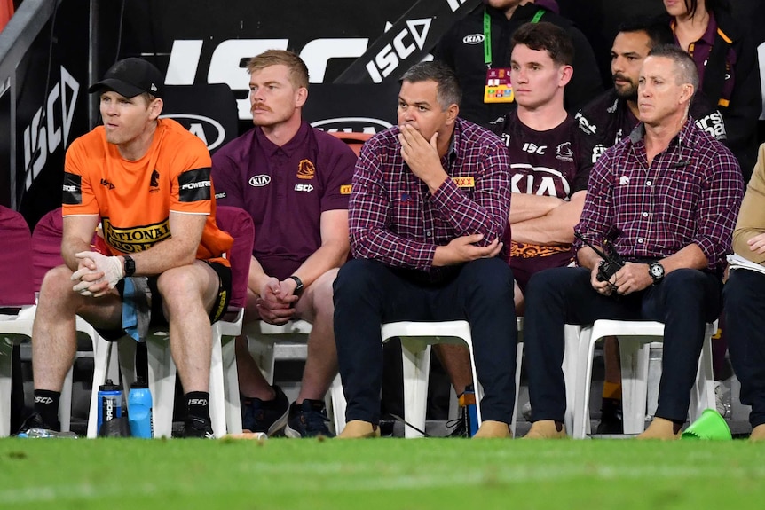 A row of young and middle-aged men sit on a bench on sideline of ground, watching a sports game.