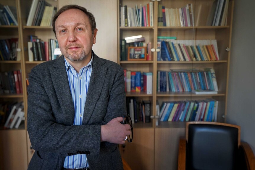 Jacek Kucharczyk wering a blazer looks at the camera with a neutral expression and his arms crossed standing near bookshelves