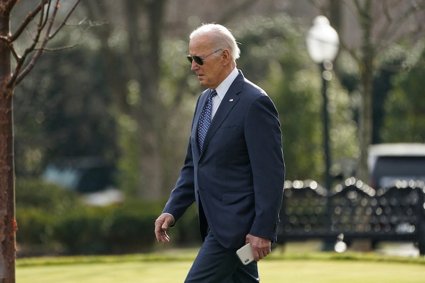 Joe Biden, in a dark blue suit, blue tie, wearing sunglasses, walking through a garden, leaving the White House