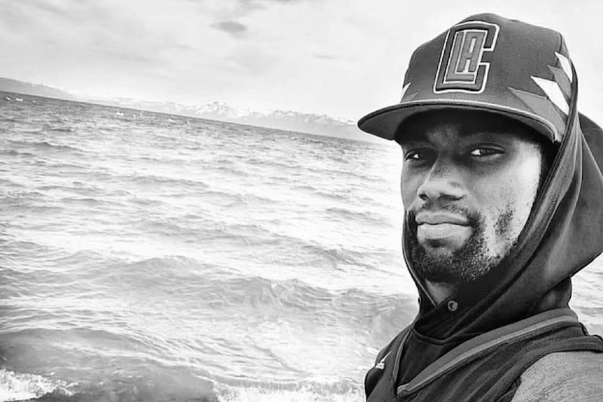 A young black man in a cap and hoodie stands at the back of a boat 