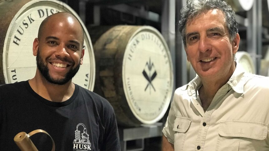 two men looking at camera with barrels of distilled spirits in the background