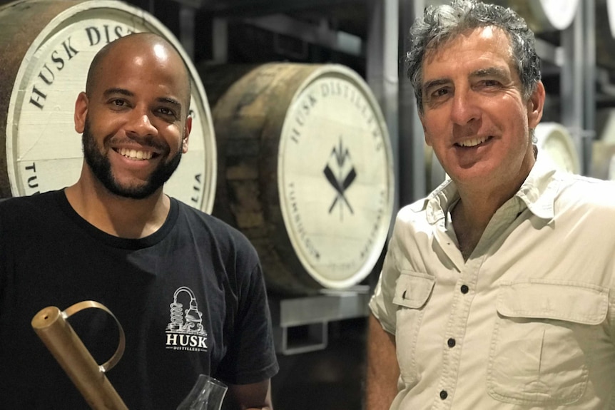 two men looking at camera with barrels of distilled spirits in the background