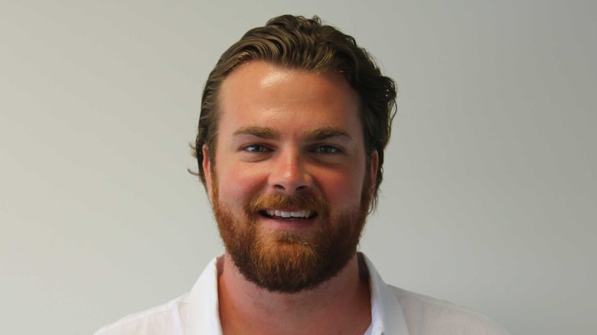 Luke Ringin wearing a white shirt and smiling at camera, in front of white wall.