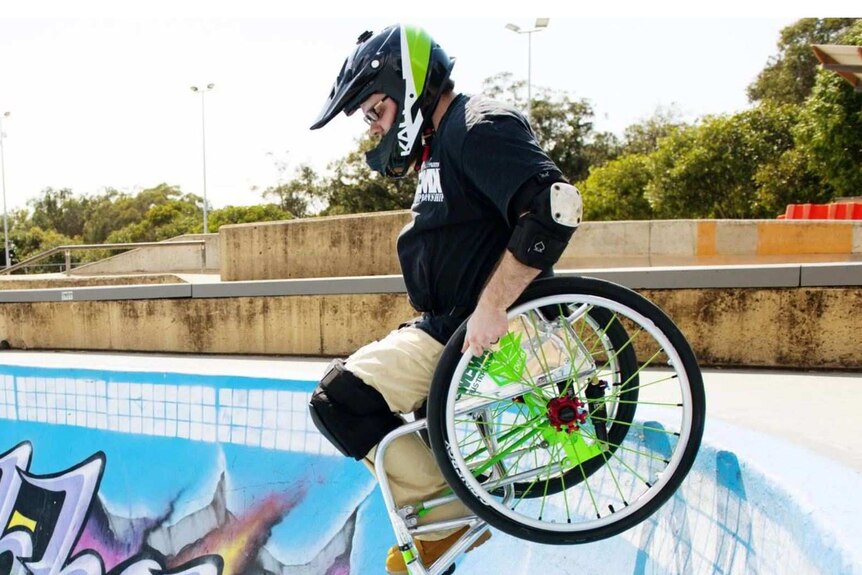 A man in a wheelchair is seen preparing to go down a dip in a skate park. He wears a helmet and knee pads.