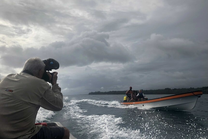 Cameraman filming boat on water with grey clouds overhead.