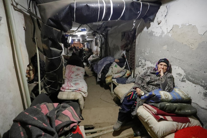 Mattresses, blankets and people line the walls of a corridor in the basement of a building. A large pipe overhead.