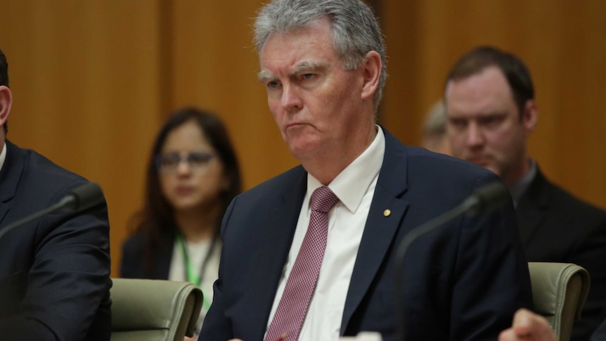Mr Lewis looks stony faced while being questioned by politicians. He has grey hair and is wearing a pink tie and navy suit.