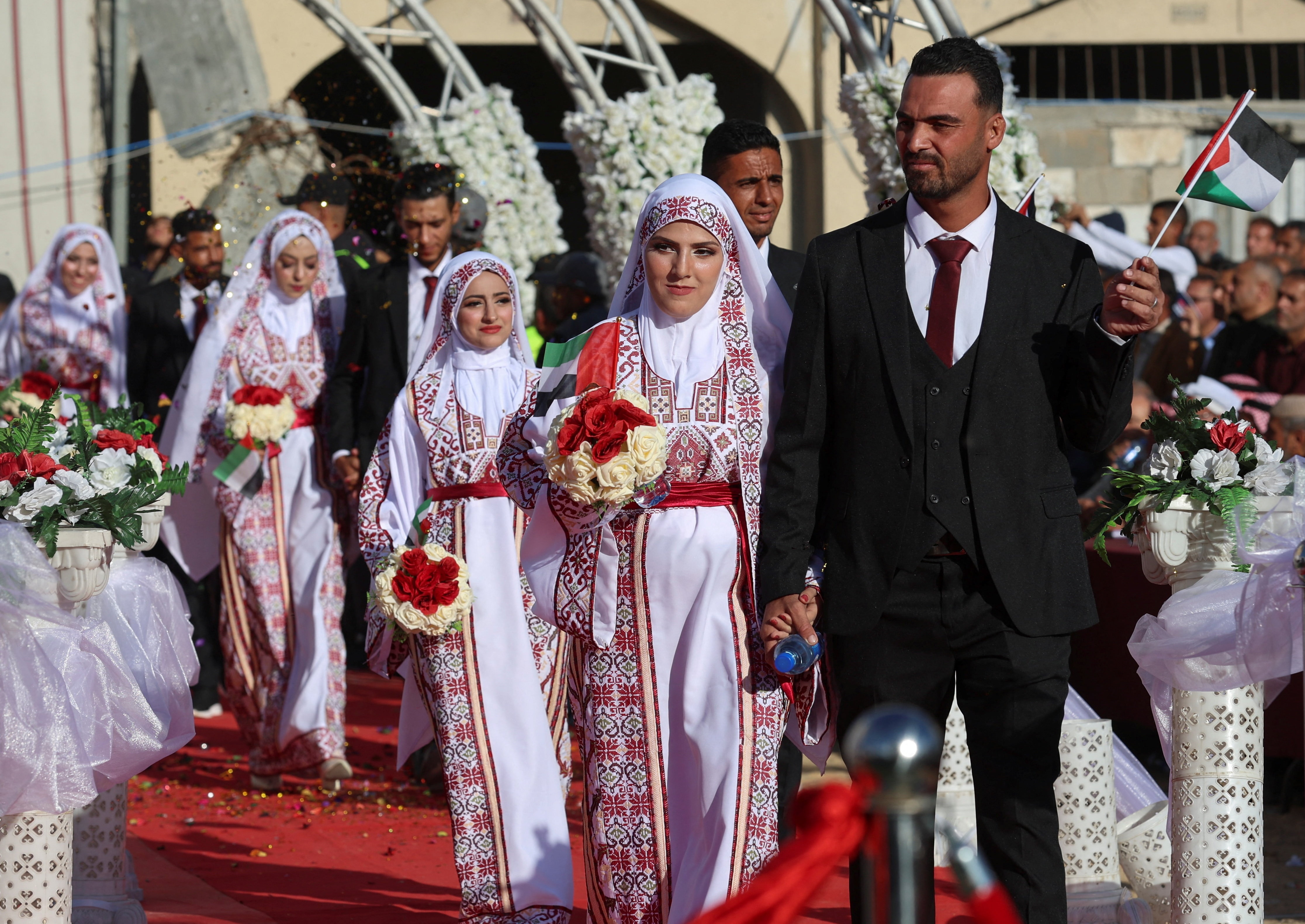 Men and women wearing suits and embroidered dresses walk on a red carpet under a flower arch.