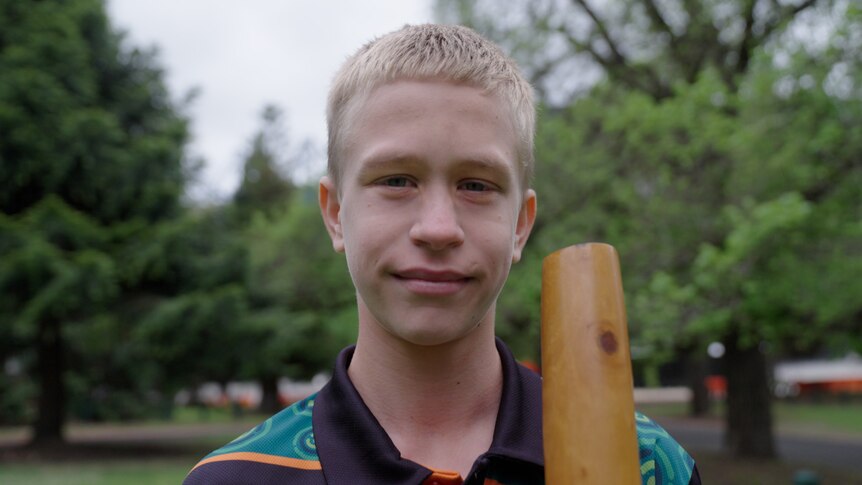boy with blonde hair smiles at camera with a didgeridoo in hand