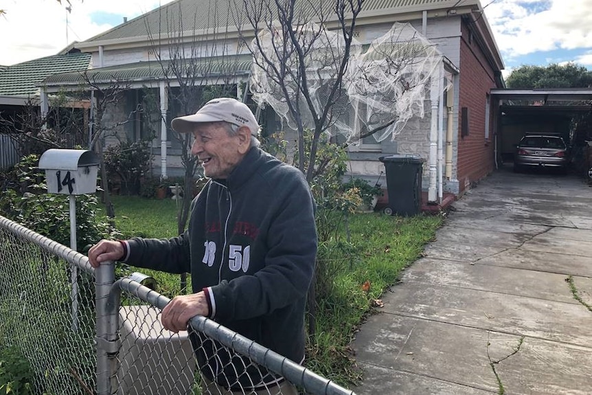 An elderly man stands in the front yard of his house leaning on the fence with trees in the background