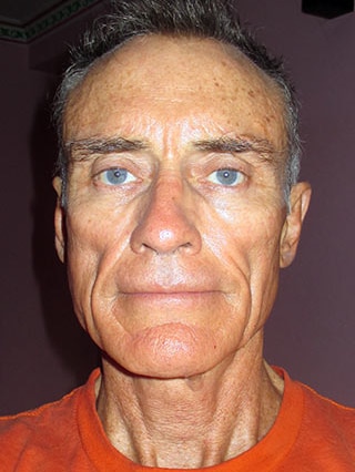 A headshot shot with flash shows a man with white hair, blue eyes and an orange shirt.