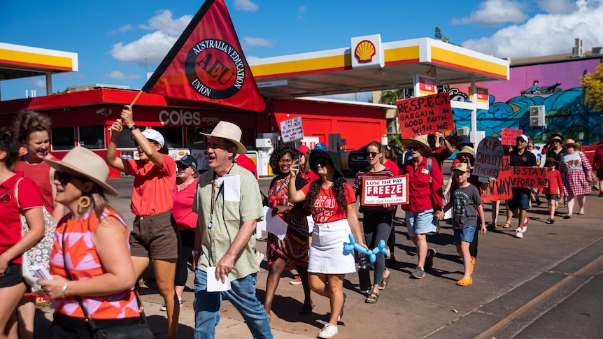 A group of people wearing mainly red clothes protesting down a street with a service station in the background