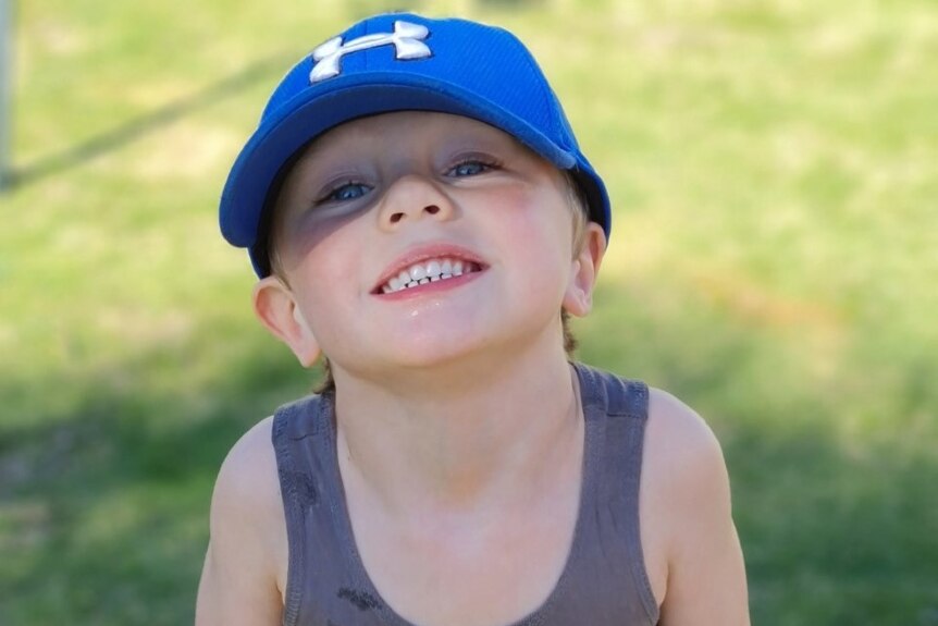 A small boy wearing a blue cap stands on a lawn.