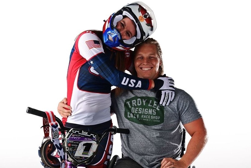A woman on a BMX bike, wearing a helmet, hugs a man in a wheelchair