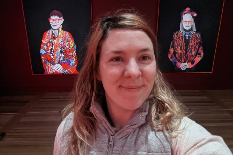 Claudia takes a selfie with two portraits of women in an art gallery.