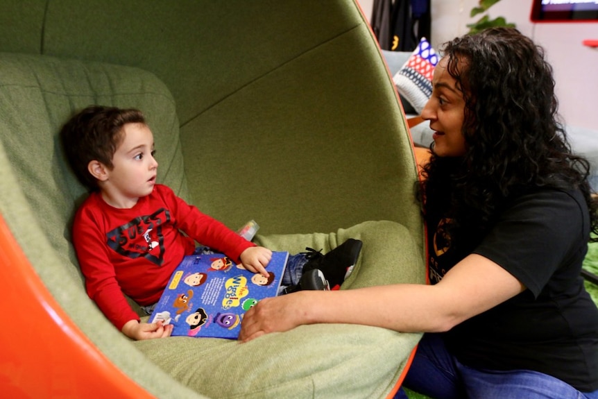 Mother looks towards toddler son sitting in chair, with an animated expression. He is pointing a his book.