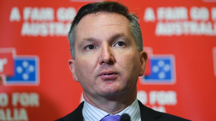 Chris Bowen wearing a suit and tie in front of a Labor party promotional backdrop.