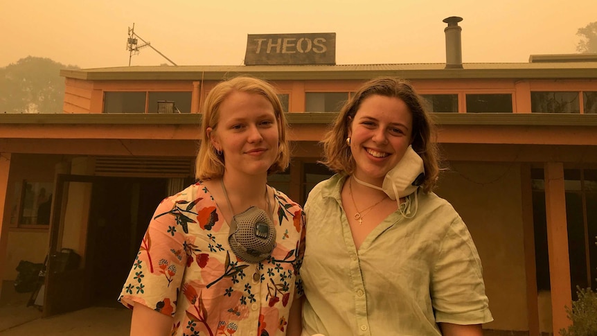 Two young woman with breathing masks around their neck smile in front of a single story building. The sky is hazy with smoke.