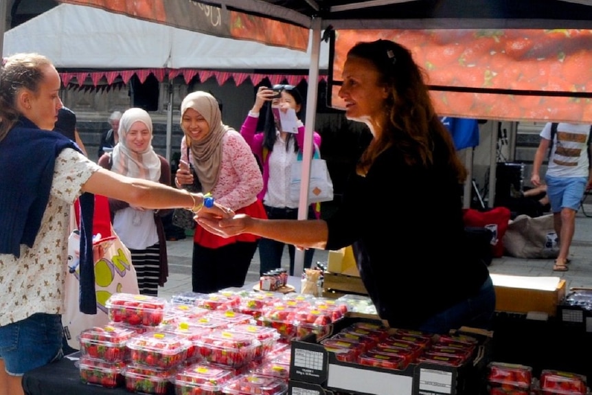 A woman hands over money to a stallholder selling strawberries as two young women look on, Melbourne University farmers market