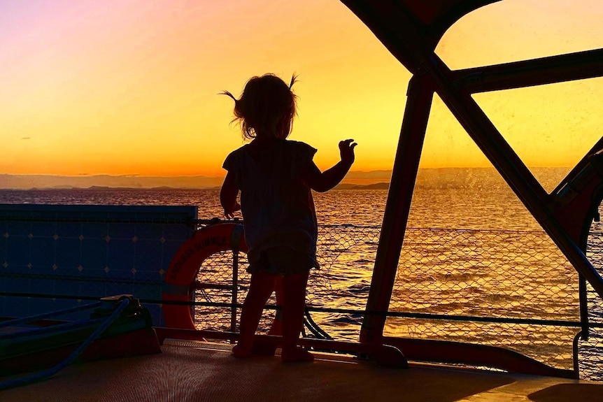 Picture of a silhouetted one-year-old girl standing on a sailboat at sunset.