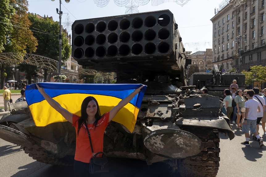 person holding ukrainian flag next to destroyed Russian tank