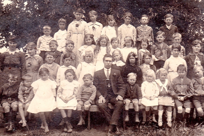 A black and white photo of a class from the early 1900s with a male teacher sitting in the middle of the students 