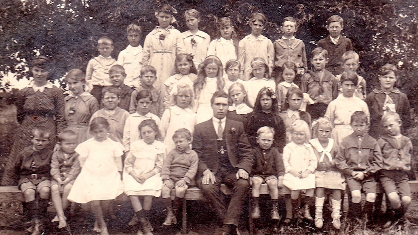 A black and white photo of a class from the early 1900s with a male teacher sitting in the middle of the students 