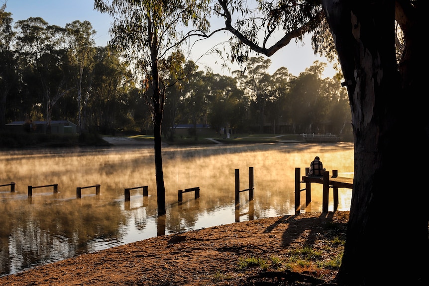 The Murray river with early morning sun on the banks and steam across the water’s surface with woman sitting on jetty