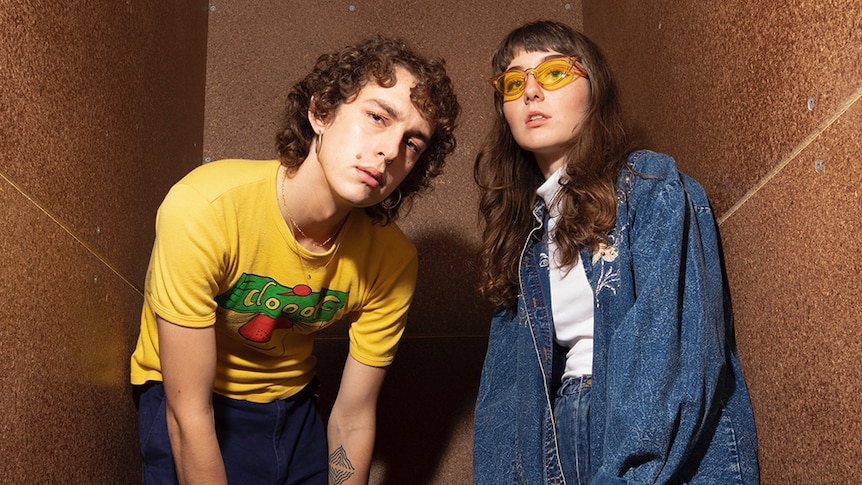 Olive (wearing a yellow t-shirt) and Scout (wearing all denim) crouch next to one another in a brown room.