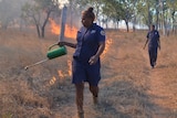 A woman using a drip torch to light fires in brown, dry open woodland.