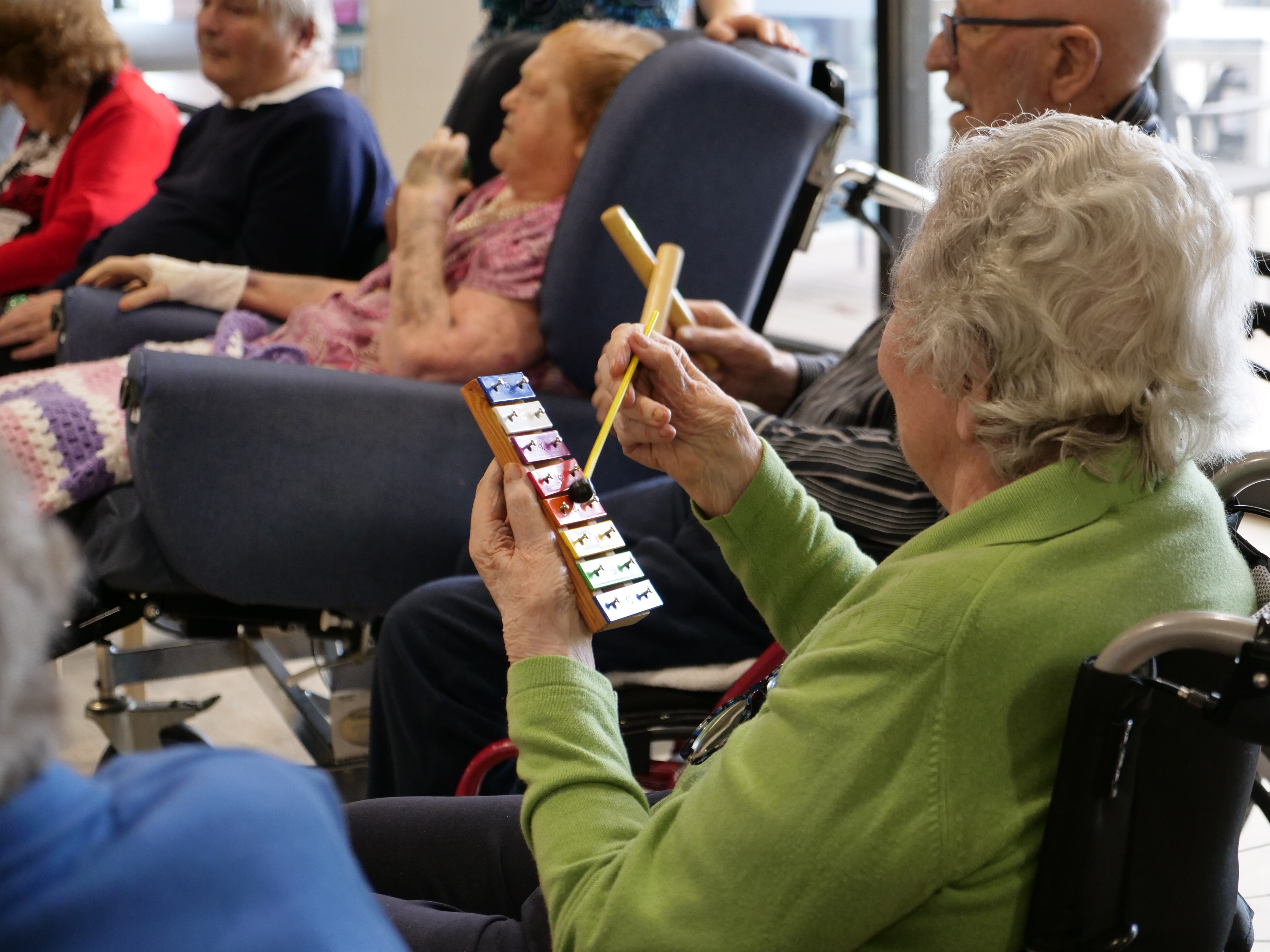 An elderly women sits in a wheelchair playing a small colourful xylophone.