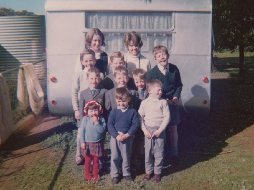 A large family of children stand smiling in front of a caravan.
