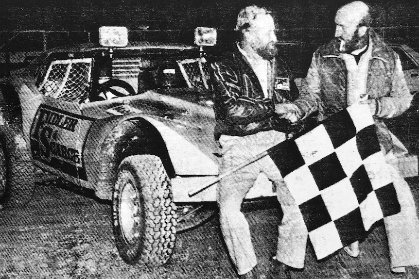 A black and white photograph of two men shaking hands in front of their car after winning the Finke Desert Race.