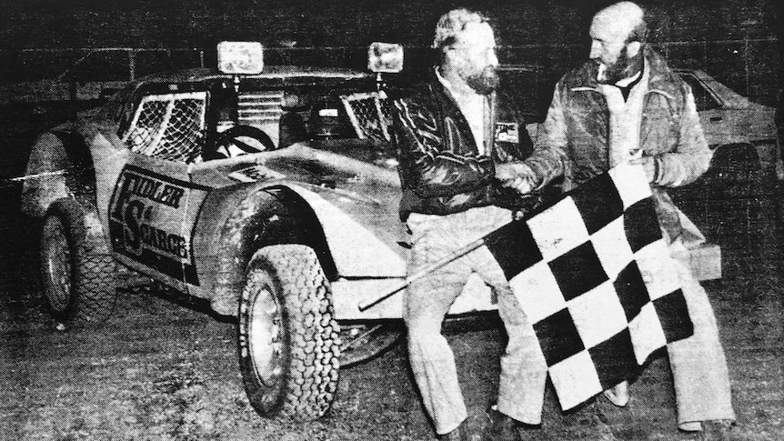 Two men shake hands in front of their car after winning the Finke Desert Race.