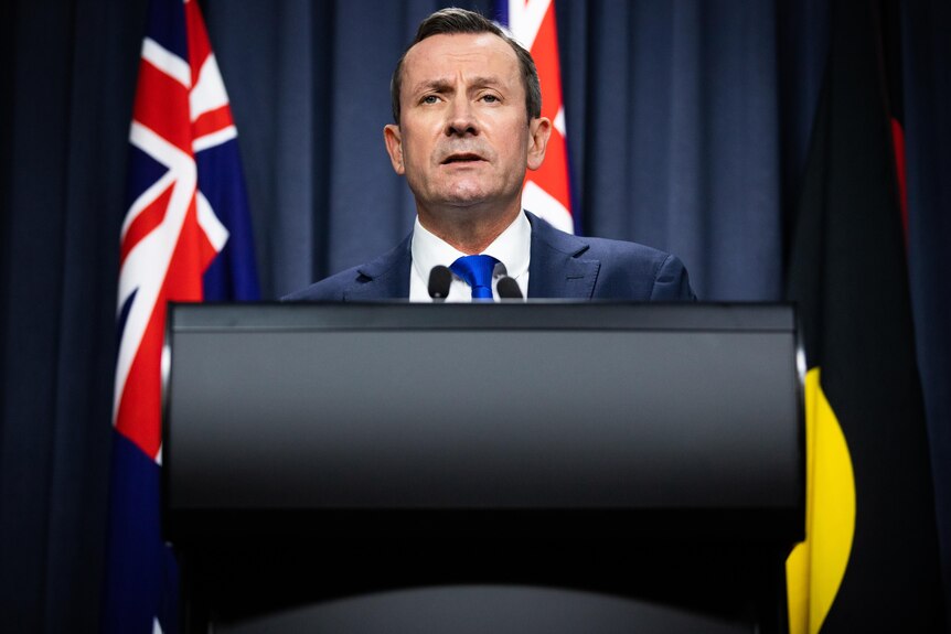 Man speaks at lectern in front of Australian, Western Australian, and Aboriginal flags.
