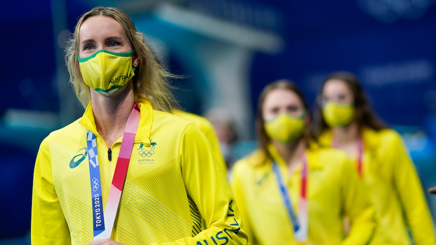 Australian swimmer Emma McKeon, wearing a green and gold mask, holds the gold medal around her neck as teammates stand behind. 