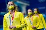 Australian swimmer Emma McKeon, wearing a green and gold mask, holds the gold medal around her neck as teammates stand behind. 