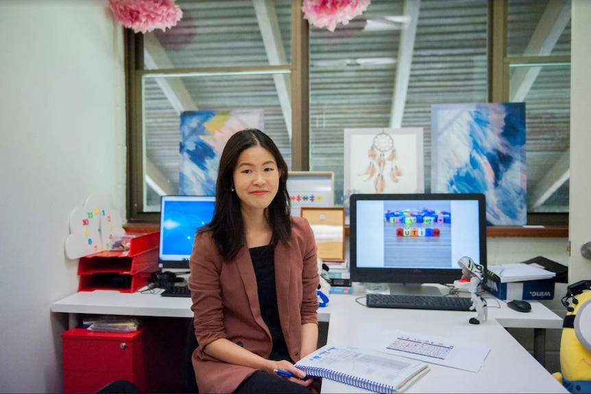 A woman with a brown jacket and black shirt sits at a desk in an office environment