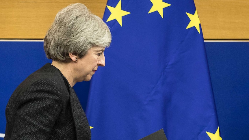 British Prime Minister Theresa May wears a dark suit and walks to the right in front of the GB and EU flags