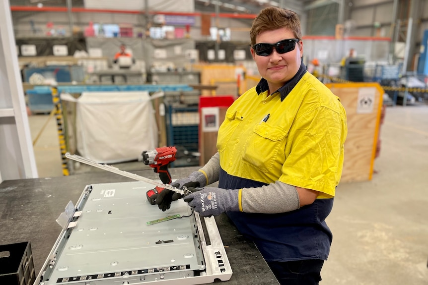 a woman in high-vis workwear and safety glasses using a drill to disassemble a television