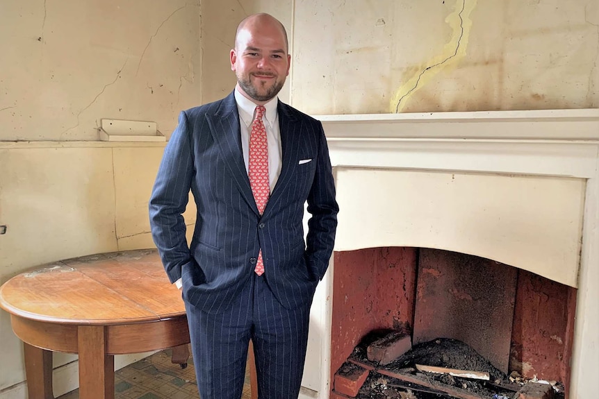 Real estate agent Brad Stephens stands in front of a fire place and table in a run down West Hobart property.