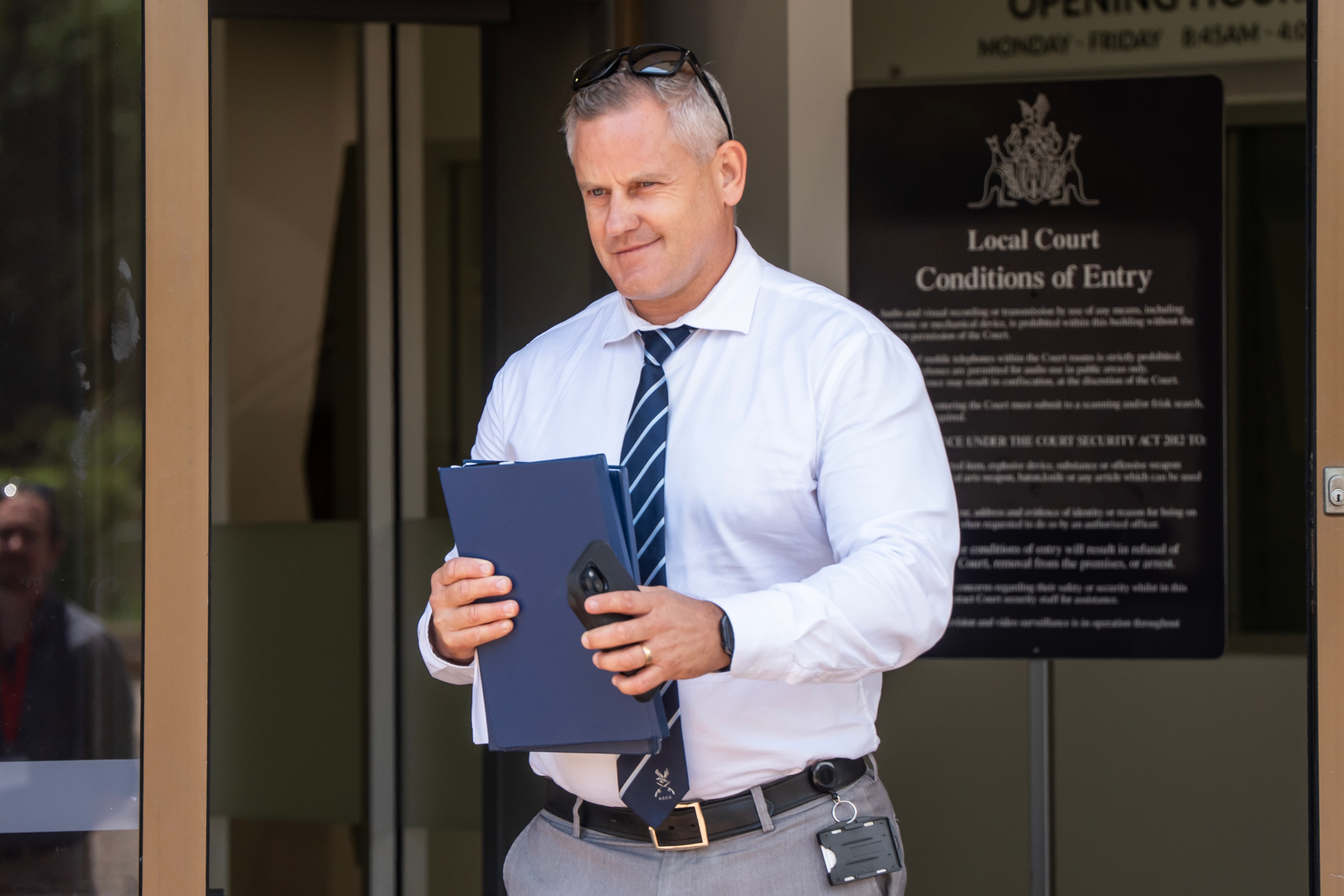 Um homem branco com camisa branca de botão, gravata listrada azul, cabelo curto e grisalho, segura pastas azuis nas mãos e sorri.