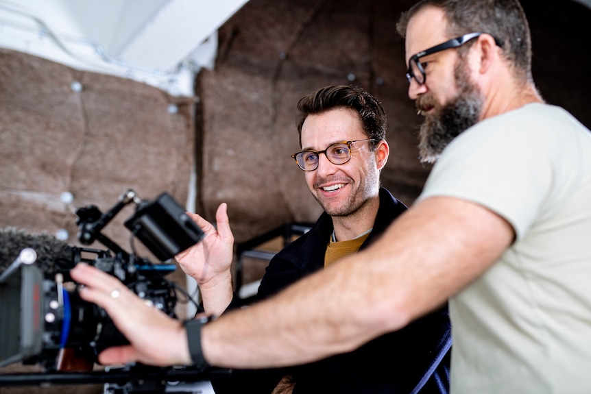 Two men smile as they look at a video camera 