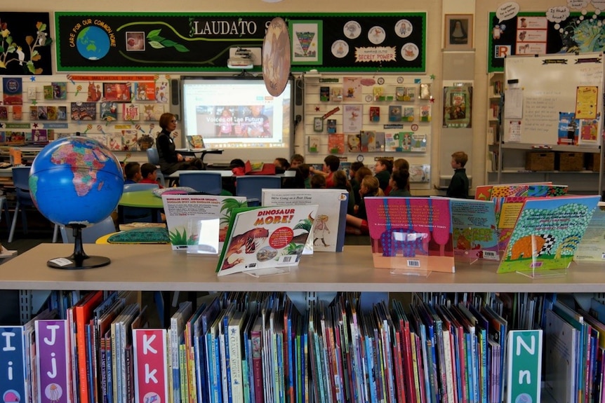 A wide interior shot of a school library with students sitting in the background on the floor.