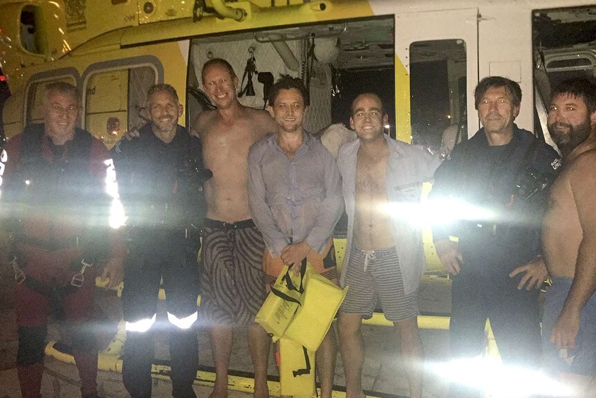 A group of men, some without shirts, link arms as they stand in front of a rescue chopper at night.