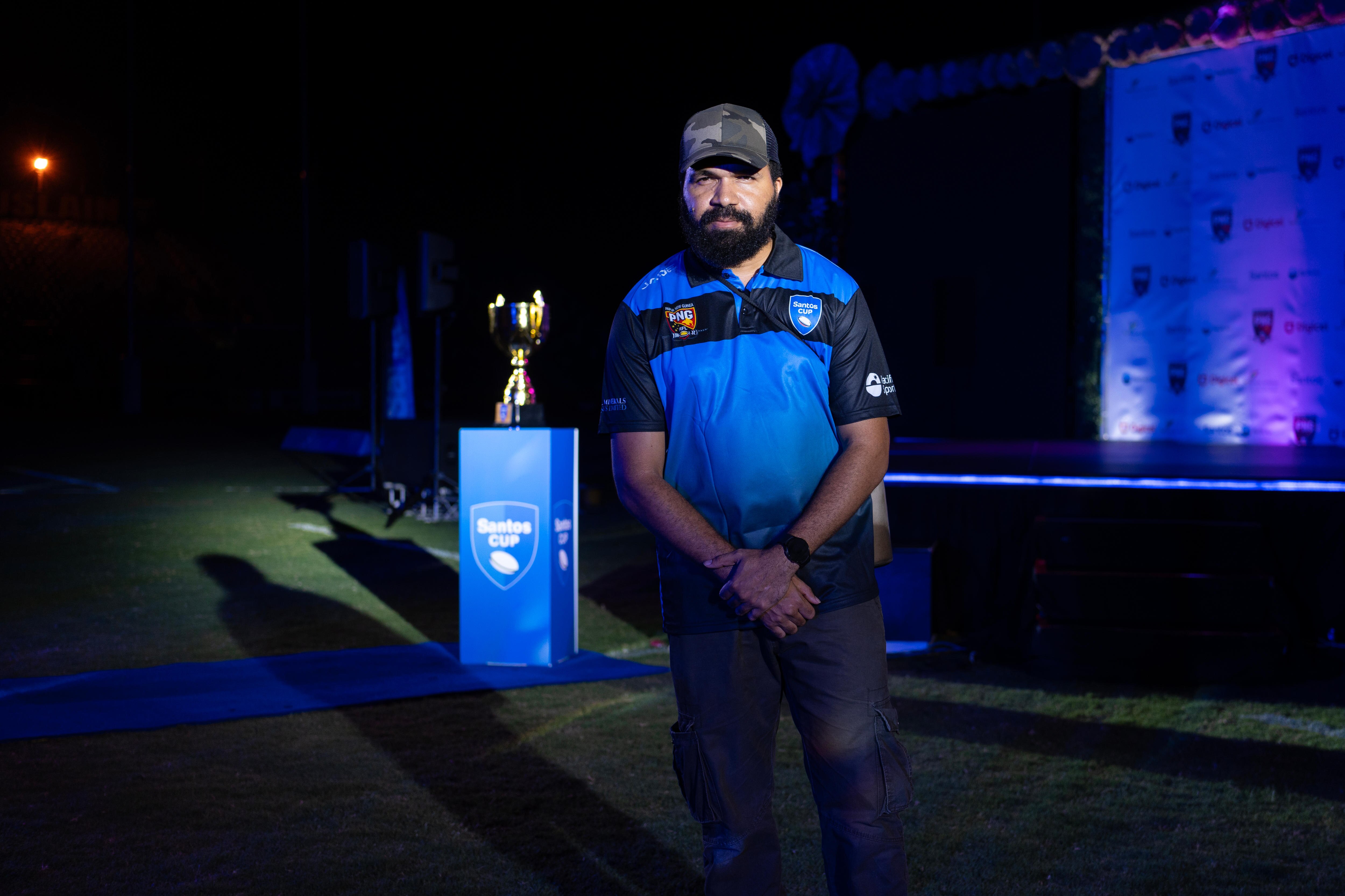 A Papua New Guinea man with a beard wearing a blue cap and blue and black shirt standing next to a trophy.