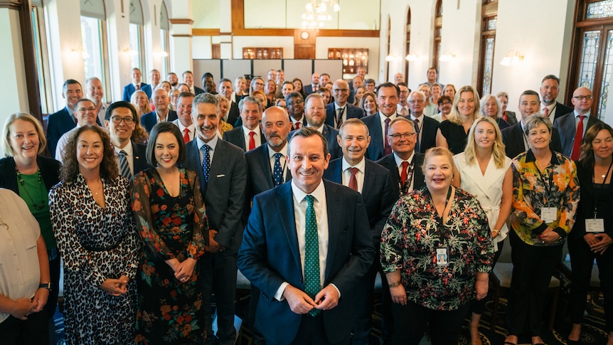 A large group of people standing up and smiling at the camera inside a room.